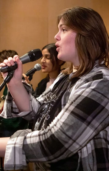 A close-up of a young singer holding a microphone and performing with a student band in a rehearsal room. A close-up of a young singer holding a microphone and performing with a student band in a rehearsal room.